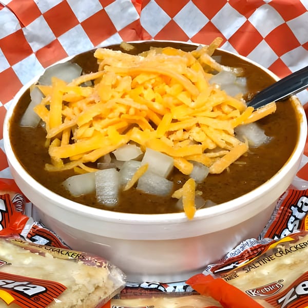 A bowl of Nicky Ds chili topped with shredded cheddar cheese and diced onions, with crackers.