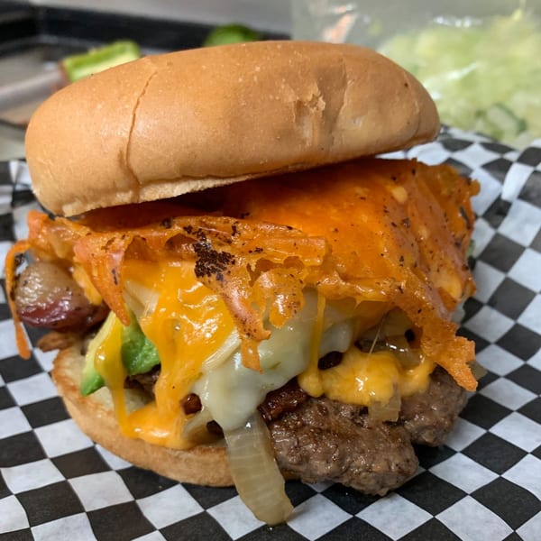 A Nicky Ds Crowley burger with melted cheese, avocado, and caramelized onions on checkered paper.